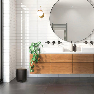 Modernes Badezimmer mit weißen Fliesen, einem schimmelfreien Holzwaschtisch mit rundem Spiegel, zwei Waschbecken und einer Wandlampe. Eine Pflanze sorgt für Leben, während der YourCasa® Mülleimer [5L] von yourcisade.com – ein schwarzer automatischer Mülleimer mit 30 % weniger Kunststoff und elegantem Design – auf dem Boden steht.
