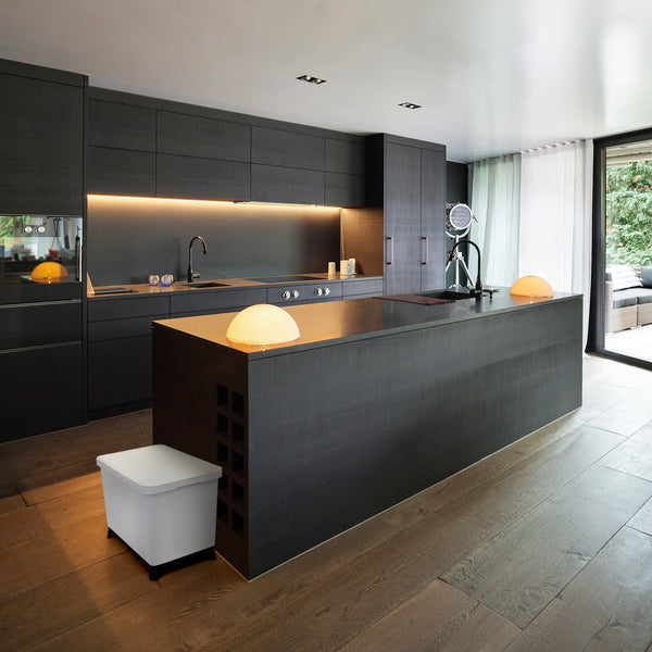Moderne Küche mit dunklen Schränken und Unterschrankbeleuchtung, mit einer zentralen Kochinsel, Hängelampen und Blick nach draußen. Ein YourCasa® Mülltrennsystem mit 2-4 Fächern [20-40L] - Effiziente Mülltrennung mit exklusivem Design ist durchdacht darin integriert.