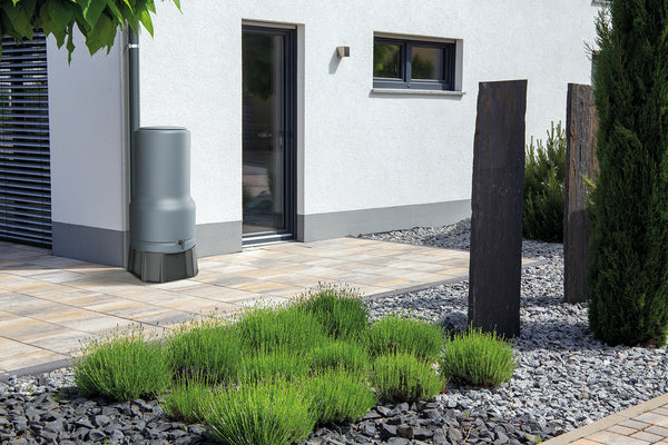 Moderne Hausaußenansicht mit einem angelegten Garten, der eine YourCasa Regentonne [RainBell] mit Standfuß, Deckel und Wasserhahn für nachhaltige Bewässerung, Ziergräser, Kies und Geometrie. Markenname: yourcisade.com