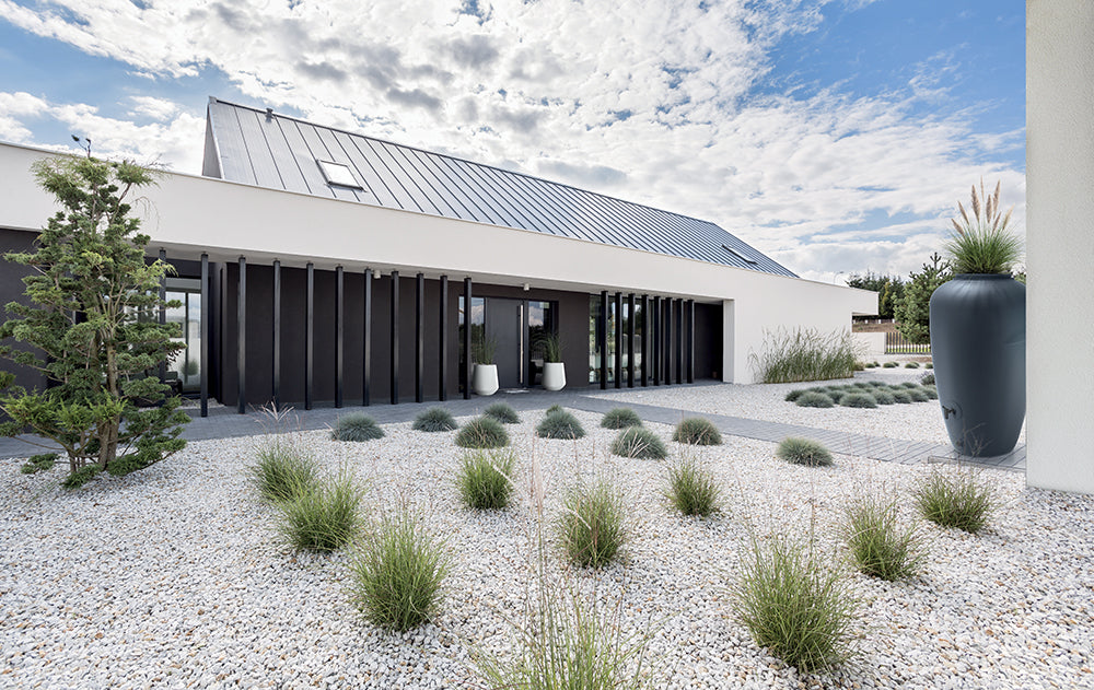 Modernes einstöckiges Haus mit einem angelegten Garten mit Ziergräsern, Kies und einer yourcisade.com Regentonne 210/360/440 Liter – Amphore Design zur Nutzung von Regenwasser.