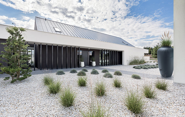 Modernes einstöckiges Haus mit einem angelegten Garten mit Ziergräsern, Kies und einer yourcisade.com Regentonne 210/360/440 Liter – Amphore Design zur Nutzung von Regenwasser.