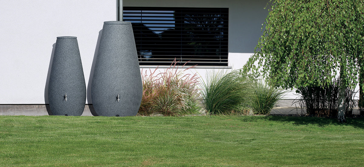 Zwei große Dekovasen neben einem modernen Gebäude mit Ziergräsern und einem Baum erleichtern die Gestaltung von YourCasa Regentonne 200 Liter [EcoDrop] – Nachhaltige Gartenbewässerung von yourcisade.com.