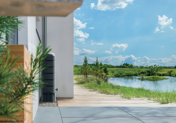 Die Außenfassade eines modernen Hauses mit Holzterrasse bietet an einem hellen, sonnigen Tag mit ein paar vereinzelten Wolken Ausblick auf eine ruhige Flusslandschaft und verfügt über eine nachhaltige Bewässerung durch eine YourCasa Regentonne [Tower] 120L mit Standfuss, Deckel und Wasserhahn von yourcisade.com.