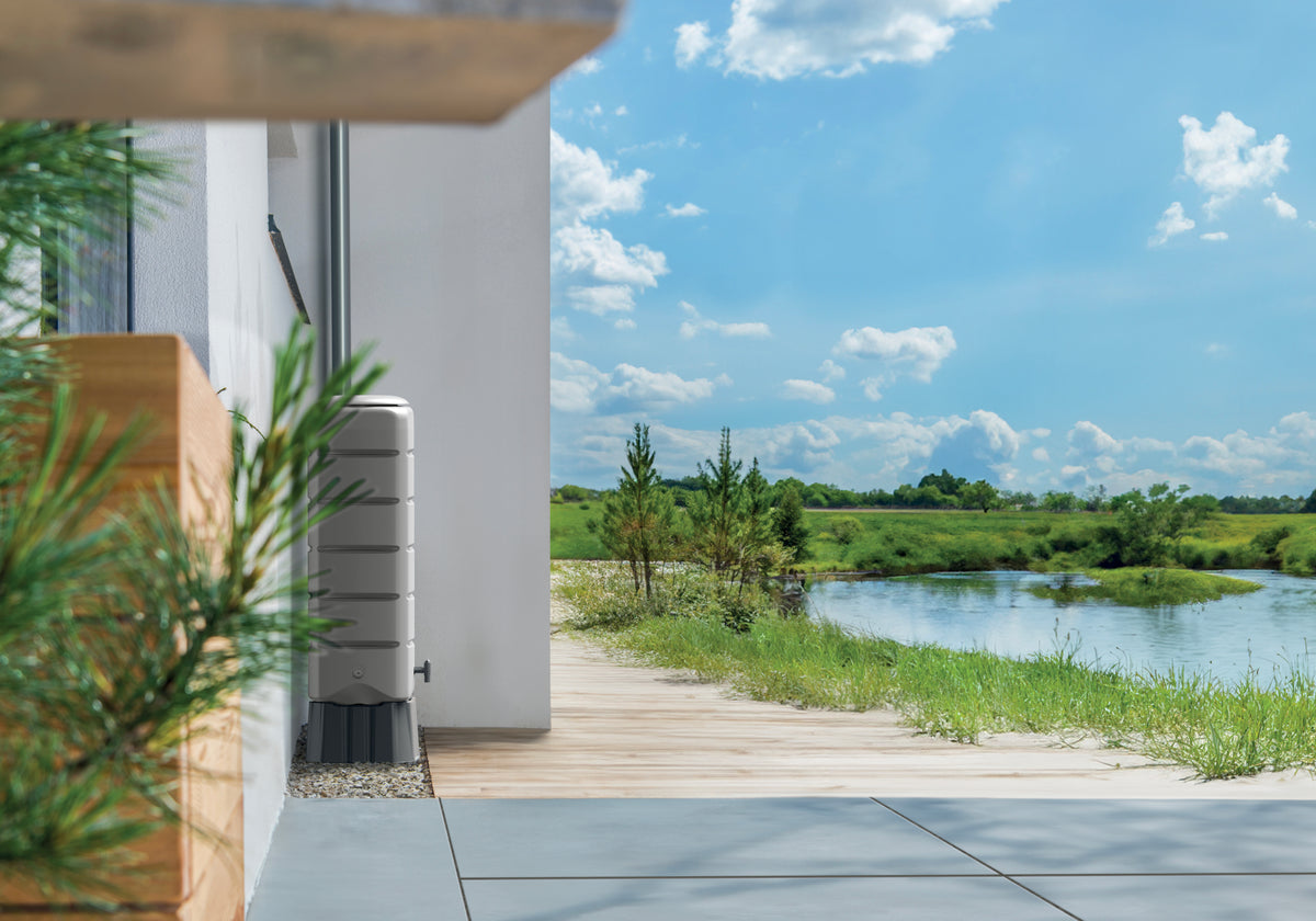 Eine moderne Hausfassade mit Holzterrasse blickt auf einen ruhigen Fluss und eine üppig grüne Landschaft unter einem blauen Himmel mit Wolken, ergänzt durch eine YourCasa Regentonne [Tower] 120L mit Standfuss, Deckel & Wasserhahn von yourcisade.com für eine nachhaltige Bewässerung.