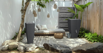 Eine minimalistische Terrasse besticht durch ein weißes Sofa, ein Holzdeck, einen Steinweg und graue Pflanzkübel, in die sich eine YourCasa Regentonne von yourcisade.com nahtlos einfügt. Sie ist frostbeständig und UV-stabil und ergänzt Hängelampen und üppiges Grün, um die ruhige Atmosphäre zu unterstreichen.