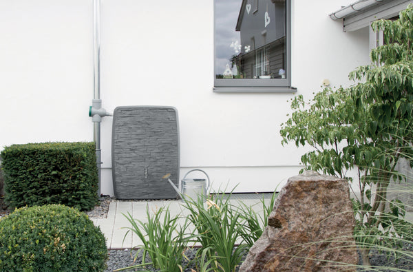 Eine YourCasa Regentonne 250 Liter [TwinView] – Eckig, schmal, platzsparend und vielseitig neben einem Haus, mit Landschaftsgestaltung mit Sträuchern und dekorativen Felsen von yourcisade.com.