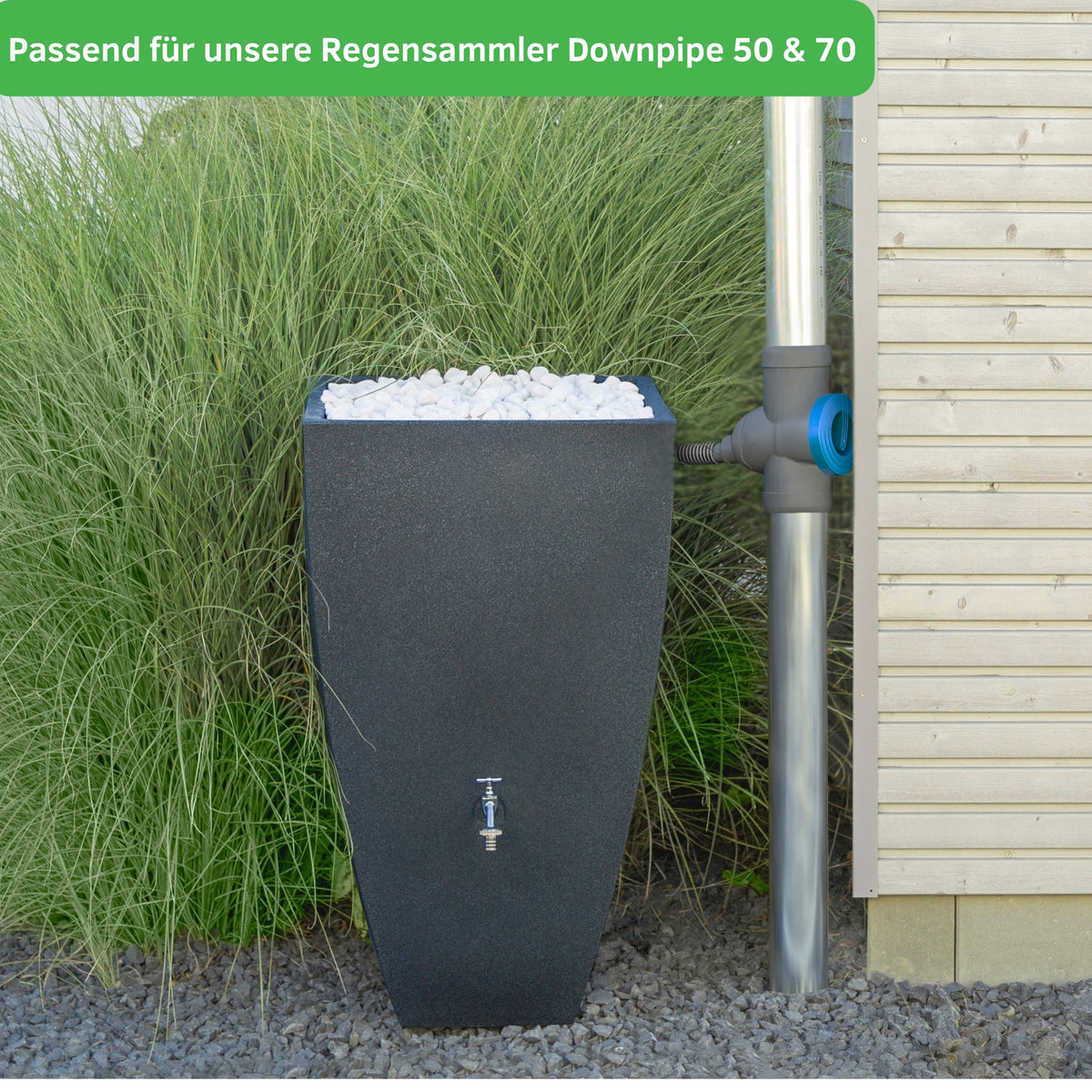 Eine hohe, schwarze Regentonne mit einem Wasserhahn ist über das yourcisade.com Regentonnen Verbinder Set 32mm inklusive Kronbohrer & Tankdurchführung mit dem Metallfallrohr neben einem Holzhaus verbunden, inmitten von hohem Gras.